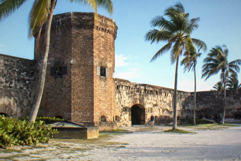 A historic brick fort exterior with a tower and palm trees under a bright sky